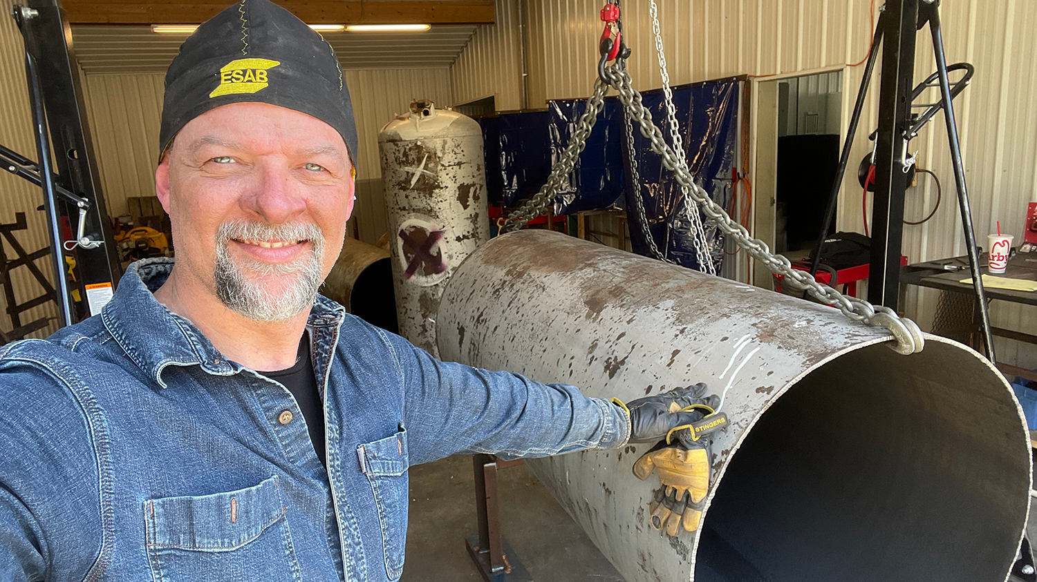 a man in welding cap and blue denim shirt standing next to a smoker being built