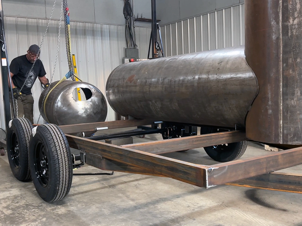 Tank smoker with a square firebox being lined up over a trailer frame in the shop